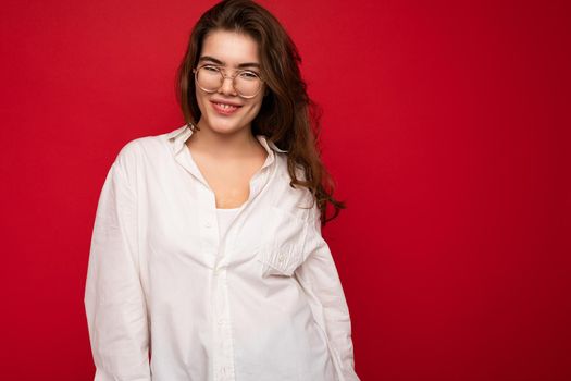 Portrait of beautiful positive cheerful cute smiling young brunette woman in casual white shirt and trendy optical glasses isolated on red background with copy space.