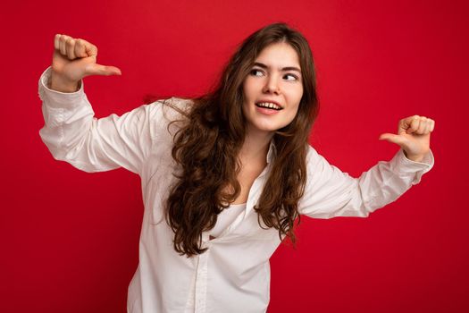 Portrait of beautiful young curly brunette woman wearing white shirt isolated on red background with free space pointing at yourself.