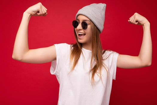 Portrait of young emotional positive happy beautiful dark blonde woman with sincere emotions wearing casual white t-shirt with empty space for mockup gray hat and sunglasses isolated over red background with free space. Female model showing powerful muscles and biceps. Fitness and workout concept.