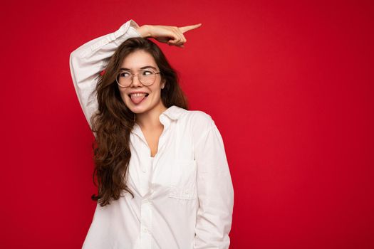 Beautiful young happy joyful funny curly brunette woman wearing white shirt and optical glasses isolated on red background with copy space and showing tongue.
