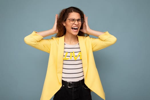 Photo of young emotional angry attractive brunette curly woman with sincere emotions wearing stylish yellow jacket and optical glasses isolated on blue background with empty space and covering ears and shouting.