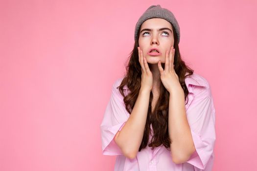 Attractive tired weary young curly brunette woman wearing pink shirt and gray hat isolated on pink background with copy space.