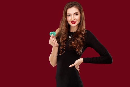 Pretty long hair woman in black dress holding chips for gambling in casino. Studio shot on red background. Poker. Two green Chips in Hand