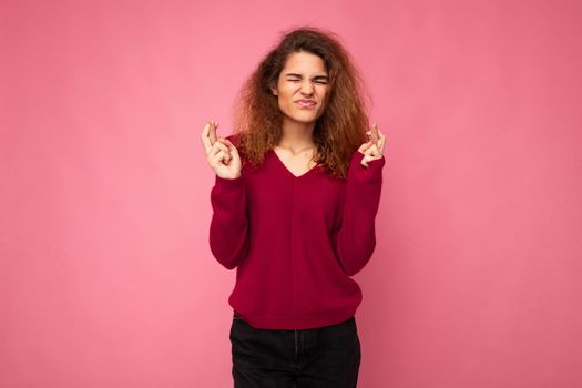 Photo shot of young beautiful brunette curly woman with sincere emotions wearing trendy pink jersey isolated over pink background with empty space and crossing fingers hoping for luck. Gesture concept.