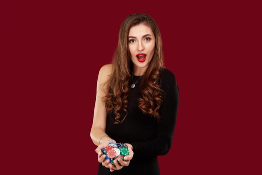 Casino, gambling, poker, people and entertainment concept - woman poker player in black dress with chips in hands on red background. Studio shot