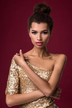 Fashion young African woman with make-up, in sexy gold dress. Model on a claret background in the studio