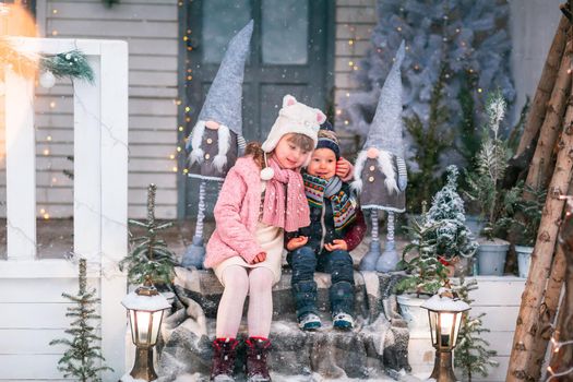 Happy little kids sitting on the porch of the Christmas decorated house, snowing outdoor. Happy New Year and Merry Christmas. Magic winter