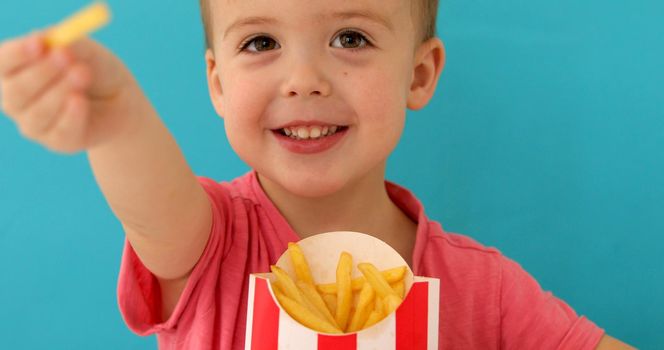 Happy cute boy smiling and offering tasty french fries to camera while eating snack against blue background
