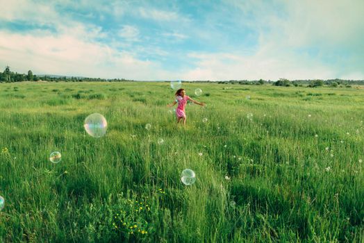 Happy little girl playing among soap bubbles on green meadow in summer