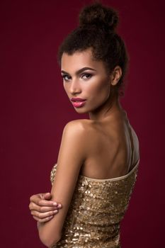 Fashion young African woman with make-up, in sexy gold dress. Model on a claret background in the studio. A woman is half a turn