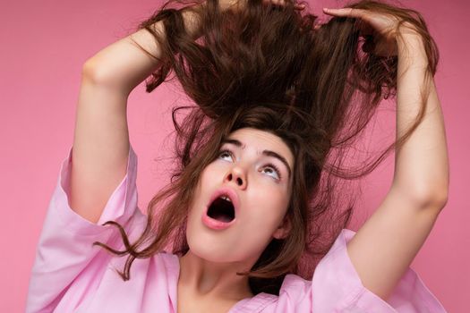 Beautiful shocked amazed young curly brunette woman wearing pink shirt and grey hat isolated on pink background with copy space.