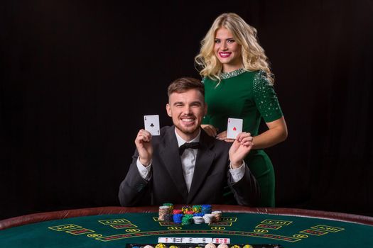 A young man in a suit shows a pair of aces. Playing poker at the table. Young woman enjoys winning at poker. They both look at the camera and smiling