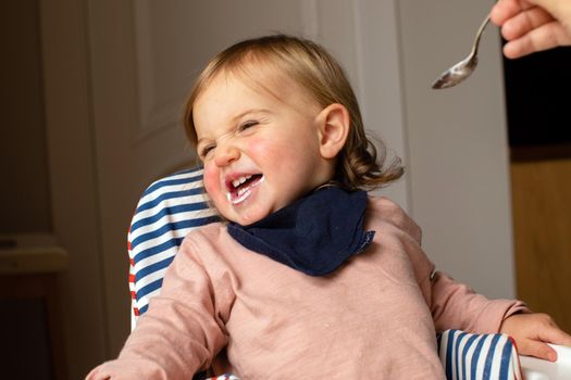 Adorable child smiling and grimacing tossing head in high chair while crop person feeding with spoon in room