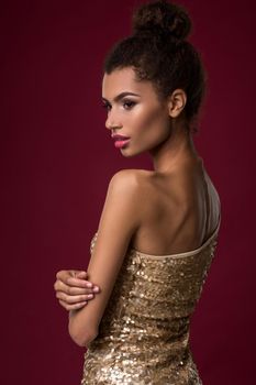 Fashion young African woman with makeup, in sexy gold dress. Model on a red background in the studio. A woman is half a turn