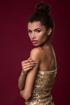 Fashion young African woman with make-up, in sexy gold dress. Model on a claret background in the studio. A woman is half a turn