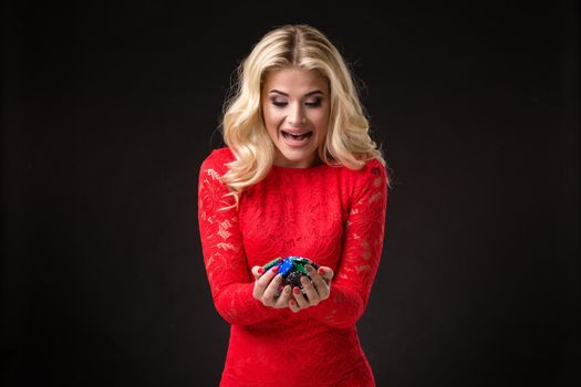 Young beautiful emotional woman with chips in hands on a black background in the studio. Portrait of a beautiful blonde in a red dress. Poker