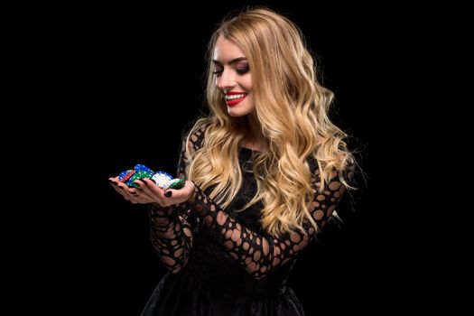Elegant blonde in a black dress, casino player holding a handful of chips on black background. Poker. Casino. Roulette Blackjack Spin. Caucasian young woman looking at the chips emotions