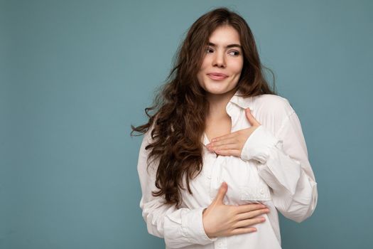 Portrait of young positive satisfied beautiful wave-haired brunette woman with sincere emotions wearing casual white shirt isolated on blue background with empty space and touching chest with hands.