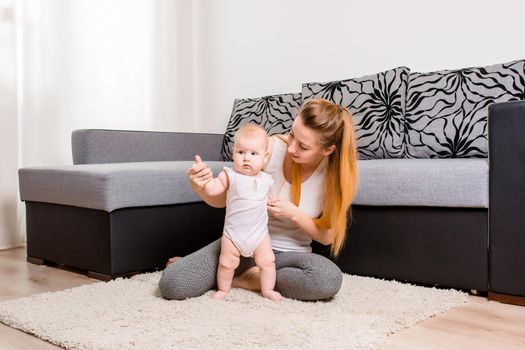 Happy young mother playing with her baby on the floor near the sofa - indoors. Mom with baby at home