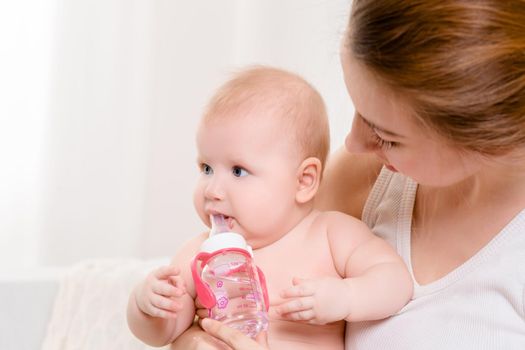 Mother and child on a white bed. Mom and baby girl in diaper playing in sunny bedroom. Parent and little kid relaxing at home. Family having fun together. Bedding and textile for infant nursery.