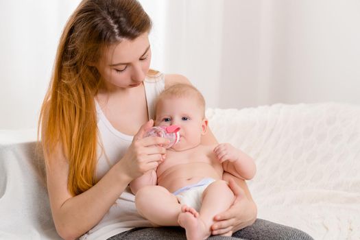 Mother and child on a white bed. Mom and baby girl in diaper playing in sunny bedroom. Parent and little kid relaxing at home. Family having fun together. Bedding and textile for infant nursery.