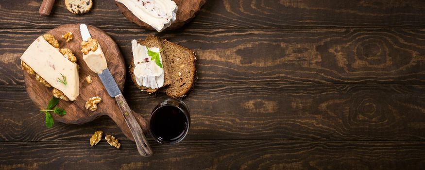 Assorted cheeses on wooden boards plate, bread and wine on dark wooden background, top view, flat lay, copy space, banner.