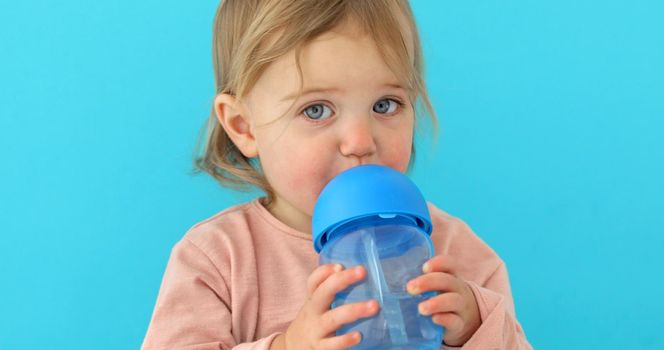 Portrait of a cute toddler drinking water from the bottle. One year old kid holding the baby cup blue background