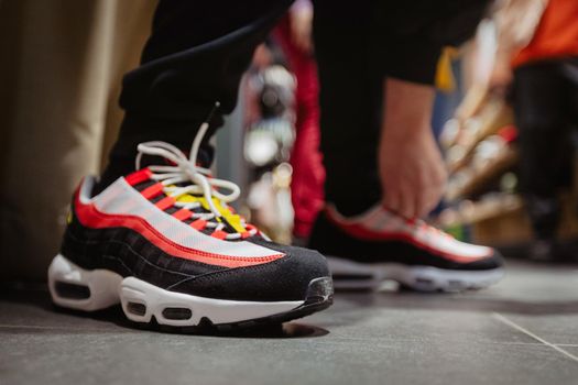 Side view of bright sneakers with black and red stripes on crop person bending to tie shoelaces on floor