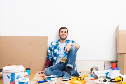 Happy smiling caucasian man relaxing on floor. Moving in new house concept. House remodeling and interior renovation. Young bearded guy sitting on floor among cardboard boxes and painting tools.