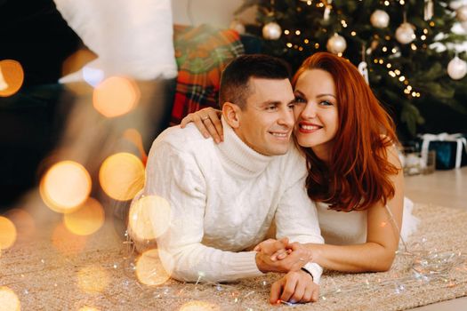 a happy married couple is lying on the floor near the Christmas tree at home.