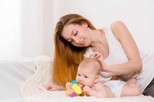 Mother and child on a white bed. Mom and baby girl in diaper playing in sunny bedroom. Parent and little kid relaxing at home. Family having fun together. Bedding and textile for infant nursery.