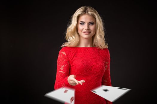 Young beautiful emotional woman throws two aces cards on a black background in the studio. Portrait of a beautiful blonde in a red dress. Poker