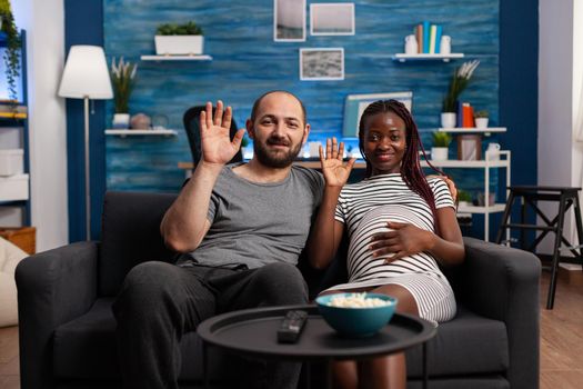 Interracial couple expecting baby and using video call for online remote communication at home. Multi ethnic parents waving while looking at camera for conference via internet connection