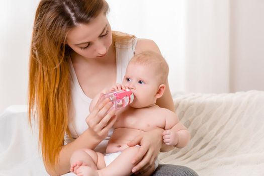 Mother and child on a white bed. Mom and baby girl in diaper playing in sunny bedroom. Parent and little kid relaxing at home. Family having fun together. Bedding and textile for infant nursery.