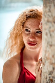 Outdoor portrait of a young beautiful natural redhead girl with freckles, long curly hair, in a red dress, posing against the background of the sea