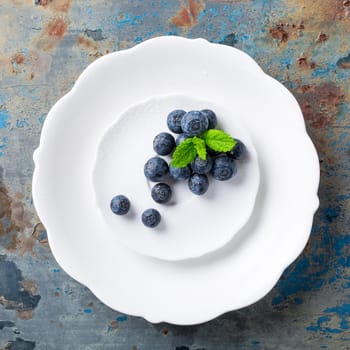 Fresh blueberries on white plate on old rusty blue background. Healthy eating and nutrition concept with copy space. Top view.