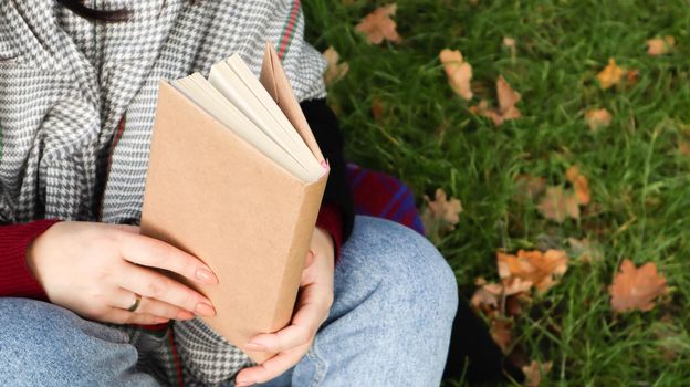 Girl reading a book in the autumn park. Female hands open the pages of a paper book outdoors on a warm sunny day. The student is preparing for the exam. Literary leisure in nature