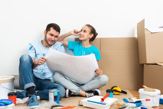 Young man and woman together planning their home renovation. Cardboard boxes, painting tools and materials on floor. House remodeling and interior renovation. People looking at blueprint at home.