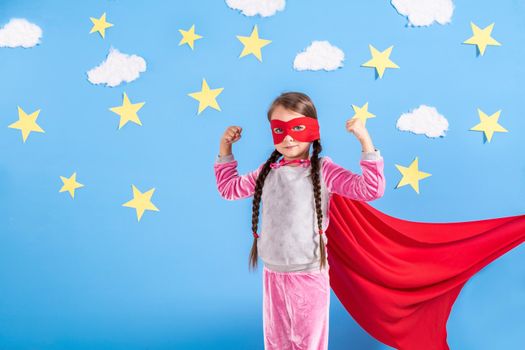 Six year blonde girl dressed like superhero having fun at home. Kid on the background of bright blue wall with white clouds and yellow stars. Girl power concept.