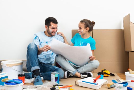 Happy couple sitting on floor with construction blueprint. Home remodeling and house interior redesign. Construction tools and materials lying on floor. Young family studies renovation project of flat