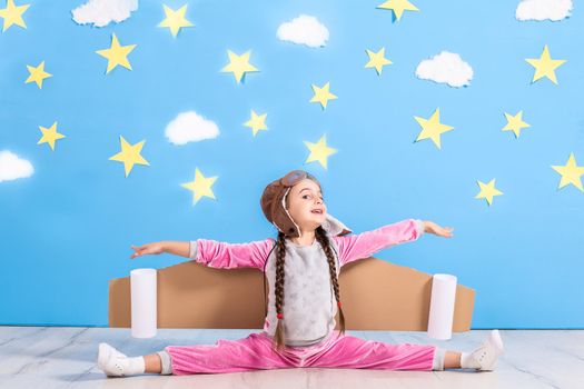 Girl pilot playing with toy jet pack at home. Success and leader concept. Kid on the background of bright blue wall with white clouds and yellow stars.