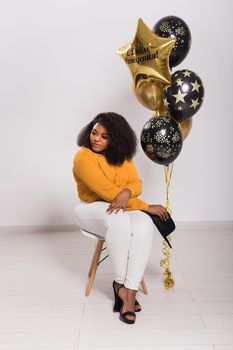 Portrait of smiling young African-American adult woman looking sweet on yellow background holding balloons.