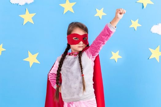 Little girl plays superhero. Kid on the background of bright blue wall with white clouds and yellow stars. Girl power concept.