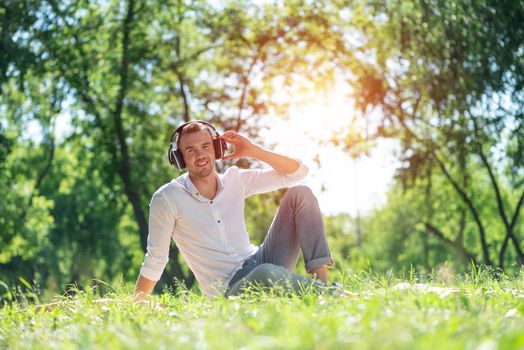 Young man in the park listens to music. He sits on the grass and enjoys music.