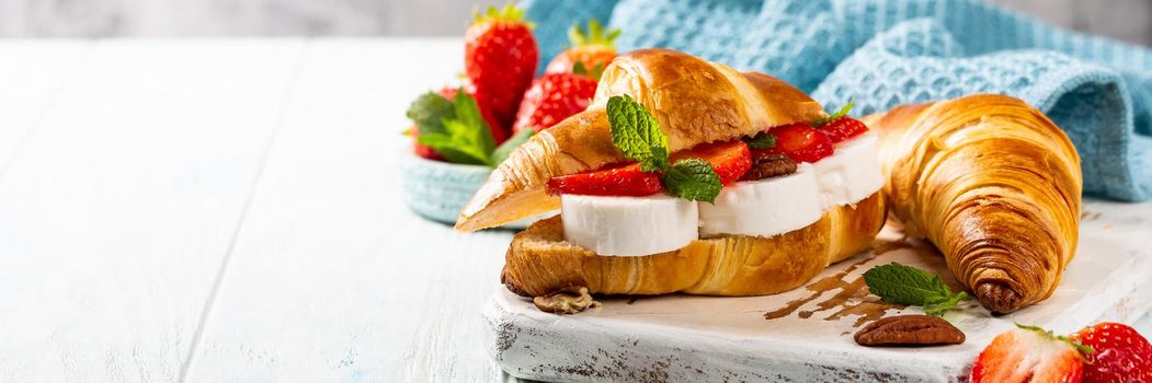 Helthy breakfast with croissant with goat cheese, strawberries, honey and pecans on wooden cutting board with copy space. Banner.