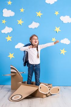 Little dreamer girl playing with a cardboard airplane at the studio with blue sky and white clouds background. Childhood. Fantasy, imagination.