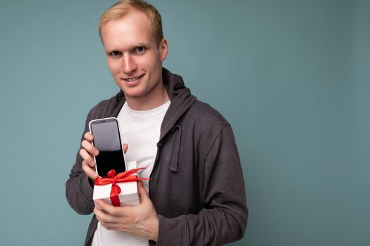 Closeup portrait photo of cool handsome happy young man wearing grey sweater and white t-shirt standing isolated over blue background wall holding smartphone and showing phone with empty screen display and gift white box with red ribbon looking at camera.