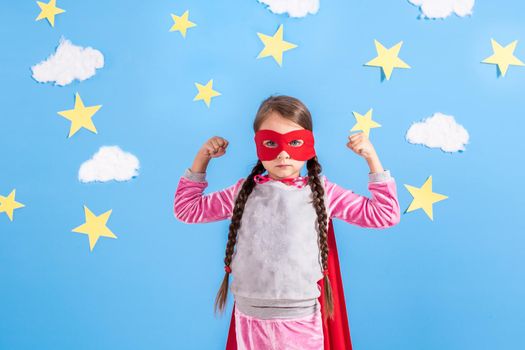 Six year blonde girl dressed like superhero having fun at home. Kid on the background of bright blue wall with white clouds and yellow stars. Girl power concept.