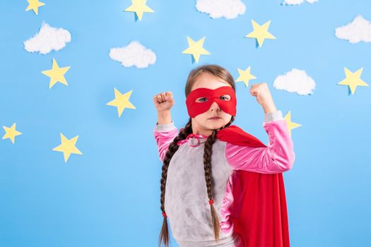 Six year blonde girl dressed like superhero having fun at home. Kid on the background of bright blue wall with white clouds and yellow stars. Girl power concept.