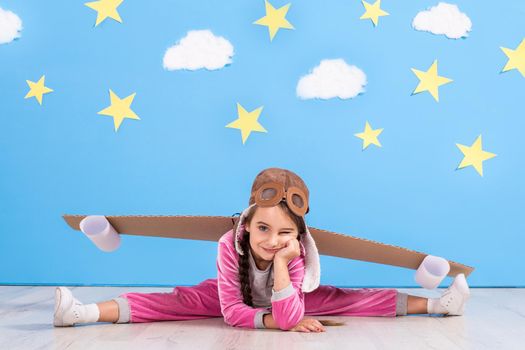 Girl pilot playing with toy jet pack at home. Success and leader concept. Kid on the background of bright blue wall with white clouds and yellow stars.